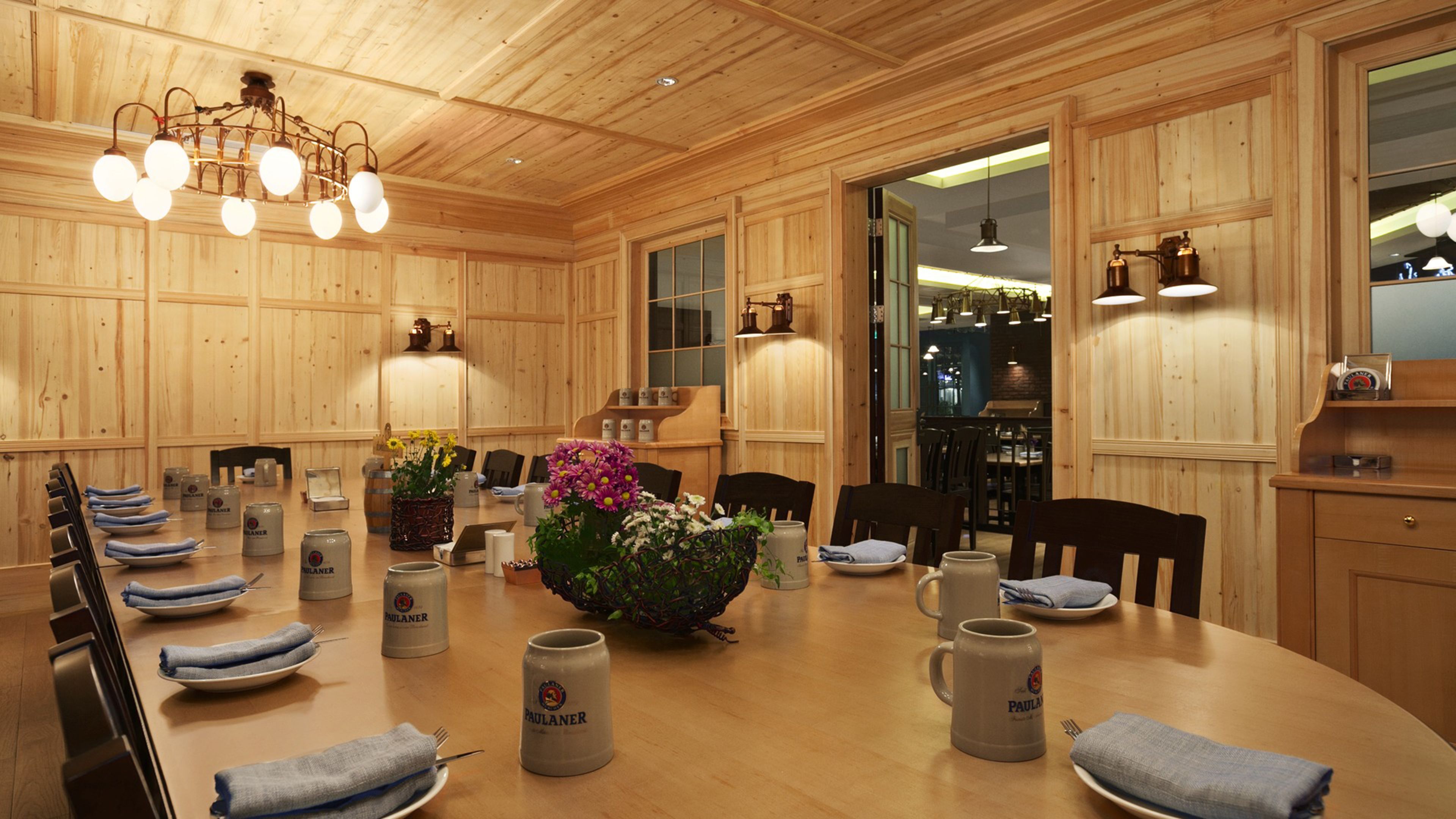 Comedor con paneles de madera y una gran mesa redonda dispuesta para una comida, con jarras de cerveza Paulaner y un centro de mesa floral.