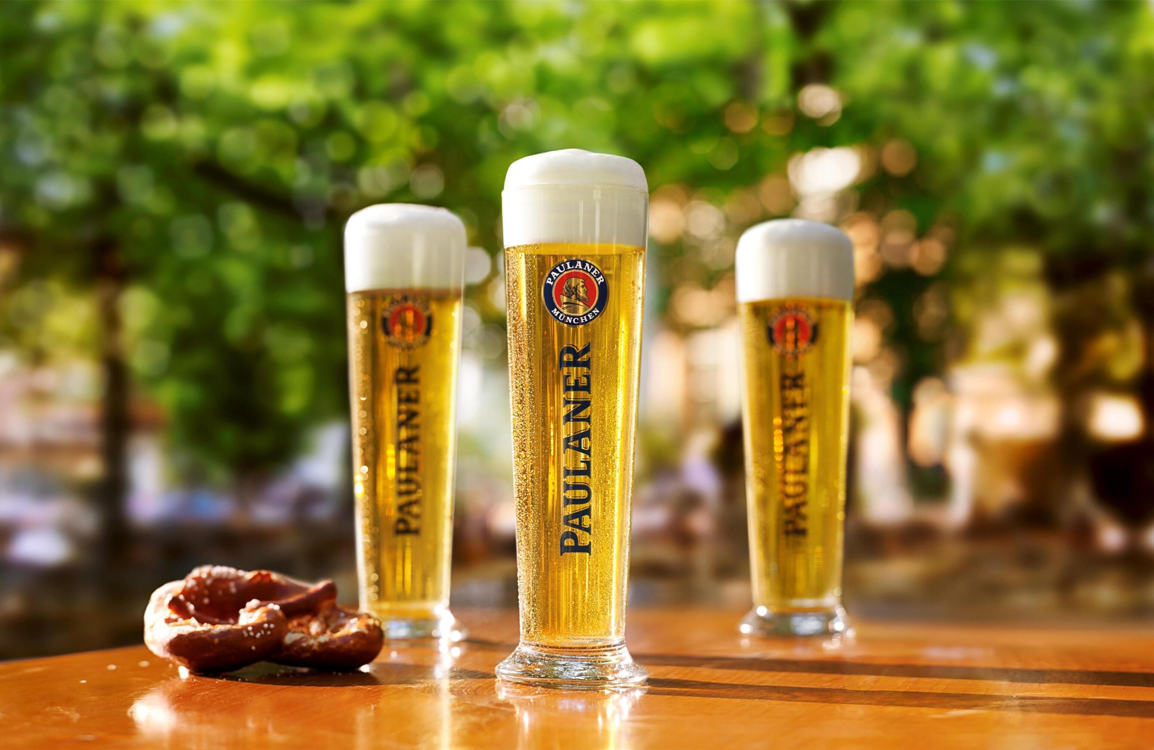 Three glasses of beer with the label "PAULANER" on a wooden surface with a pretzel.
