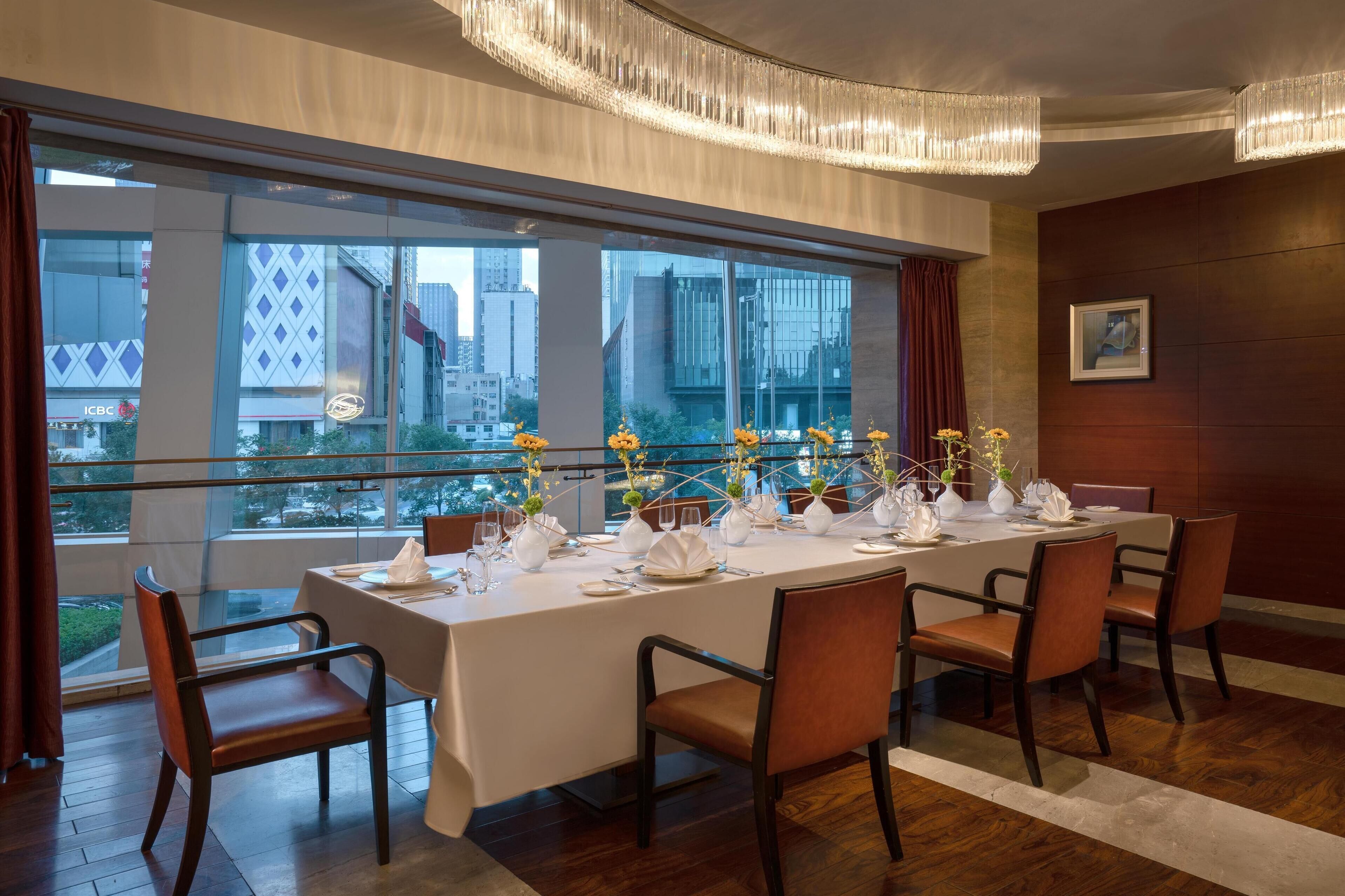 A dining area with a long table set for six, large windows, and modern chandeliers.