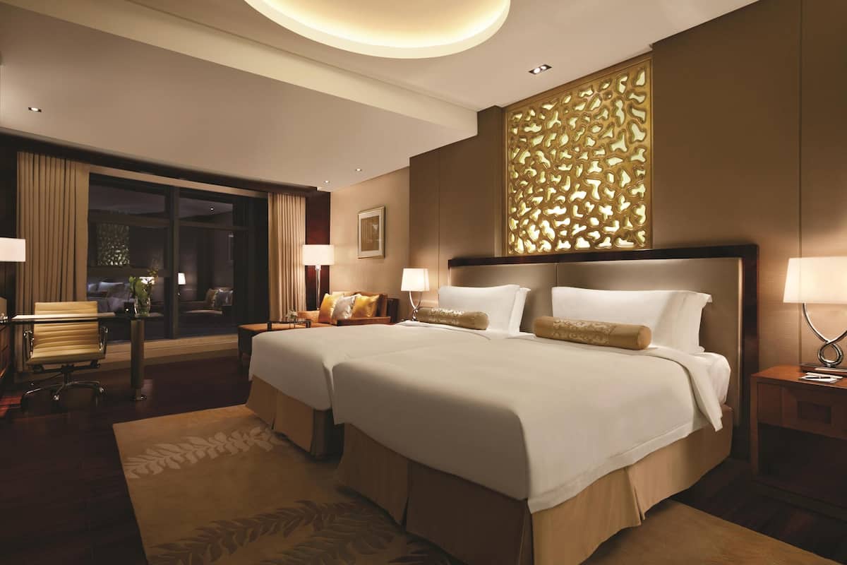Une chambre d’hôtel avec deux lits, un coin salon comprenant une chaise et une table, une grande fenêtre et un panneau décoratif au mur.