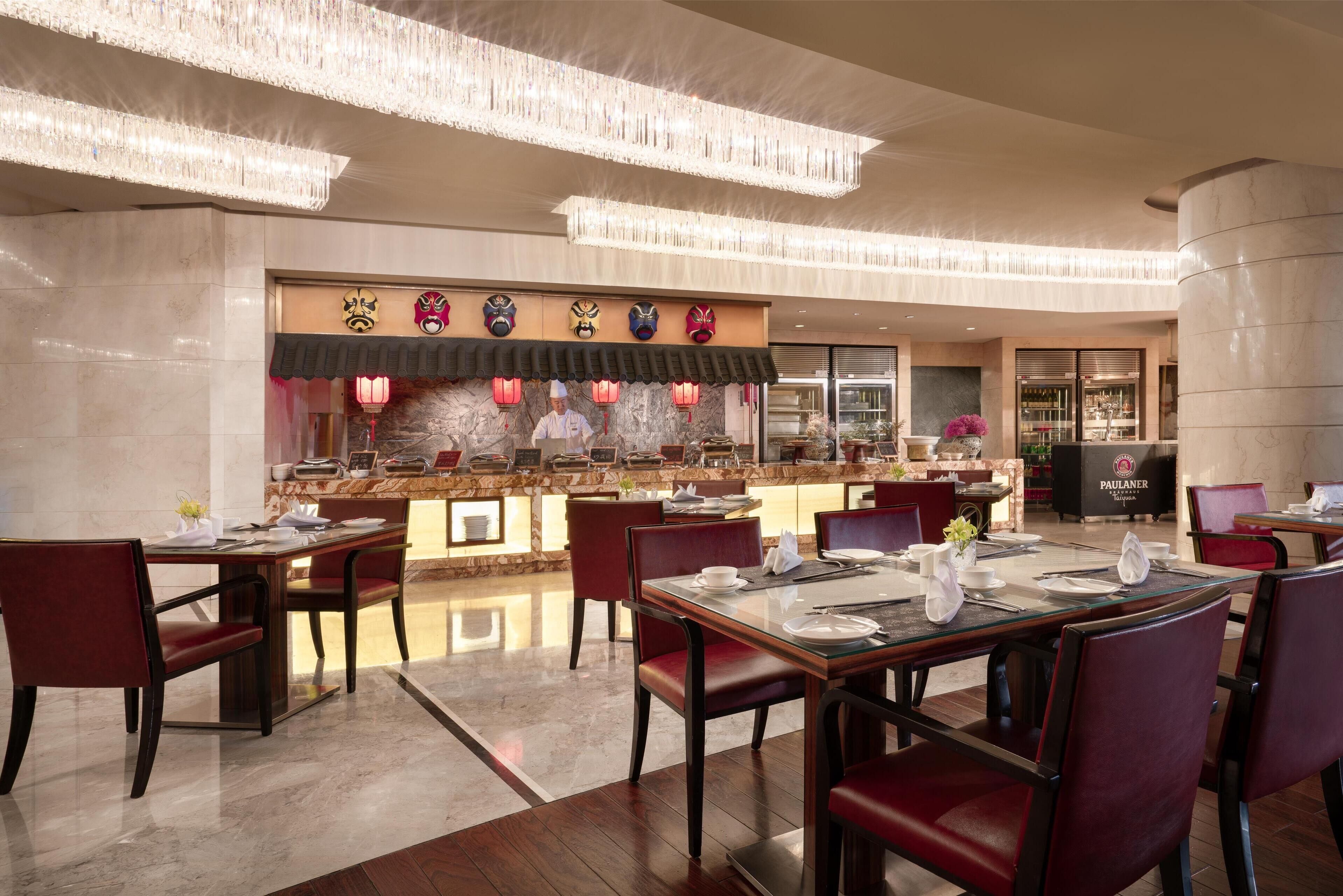 A restaurant with a traditional Asian theme, featuring a counter with various items and a display of masks. Tables are set with red chairs and white tableware.