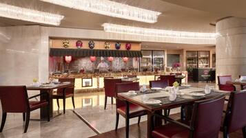 A restaurant with a traditional Asian theme, featuring a counter with various items and a display of masks. Tables are set with red chairs and white tableware.