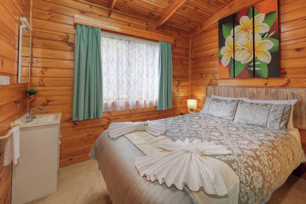 Une chambre à coucher dans une cabane en bois, avec un lit, une table de chevet, une lampe, un lavabo et une fenêtre avec des rideaux.