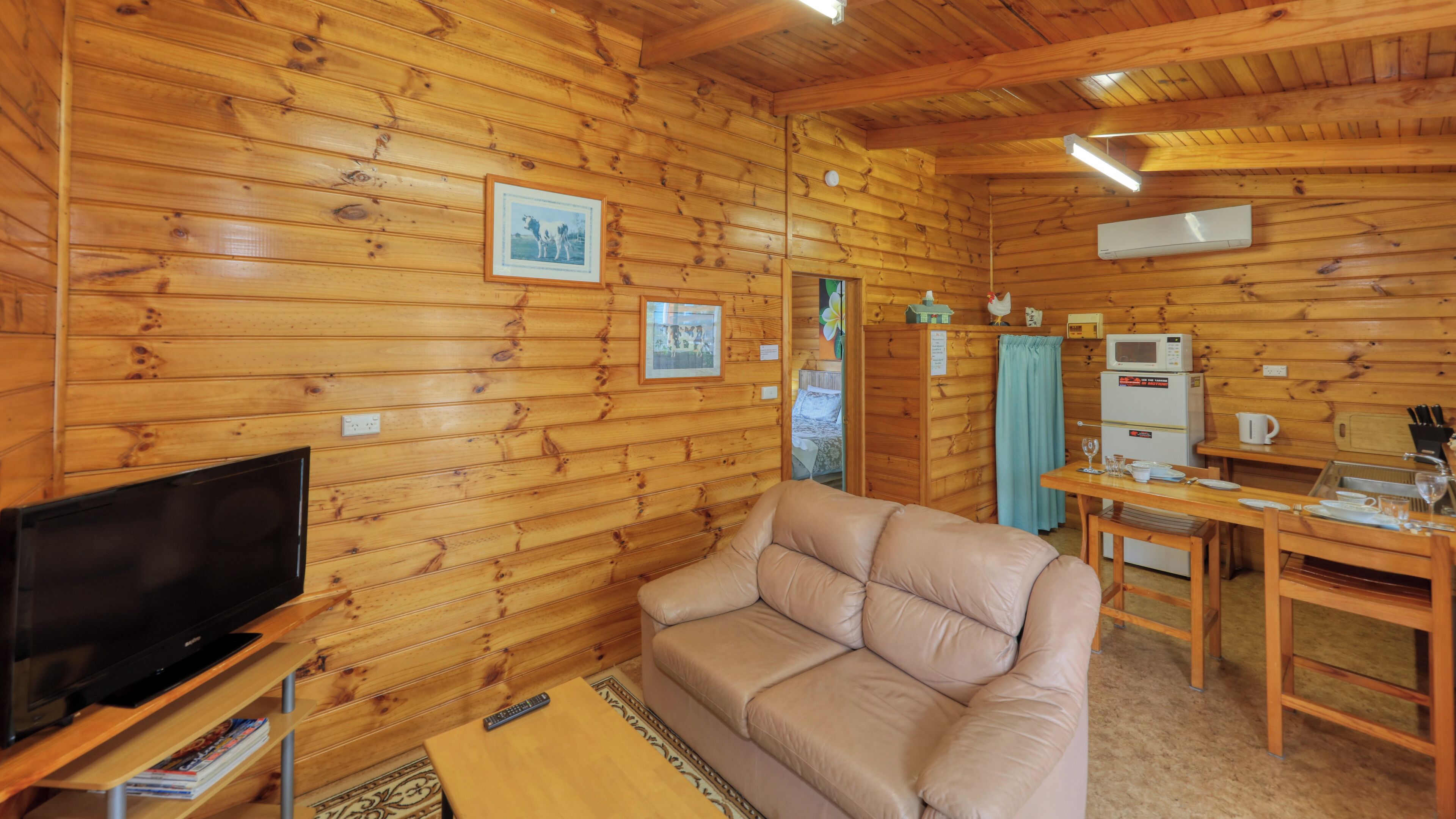 Un intérieur de chalet chaleureux avec un canapé en cuir, un téléviseur à écran plat et une kitchenette équipée d’un four à micro-ondes.