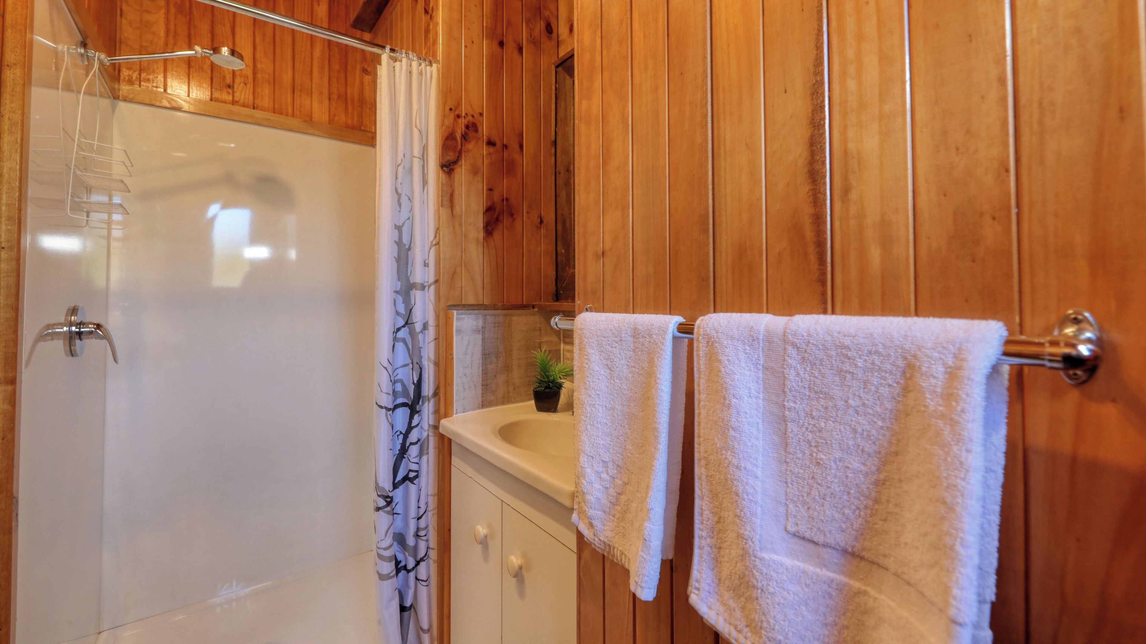 Un baño de madera con ducha, lavamanos y dos toallas colgadas en un perchero.