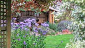 Une construction rustique en bois, dotée d’un toit de chaume, entourée d’un jardin luxuriant où poussent diverses plantes et fleurs.