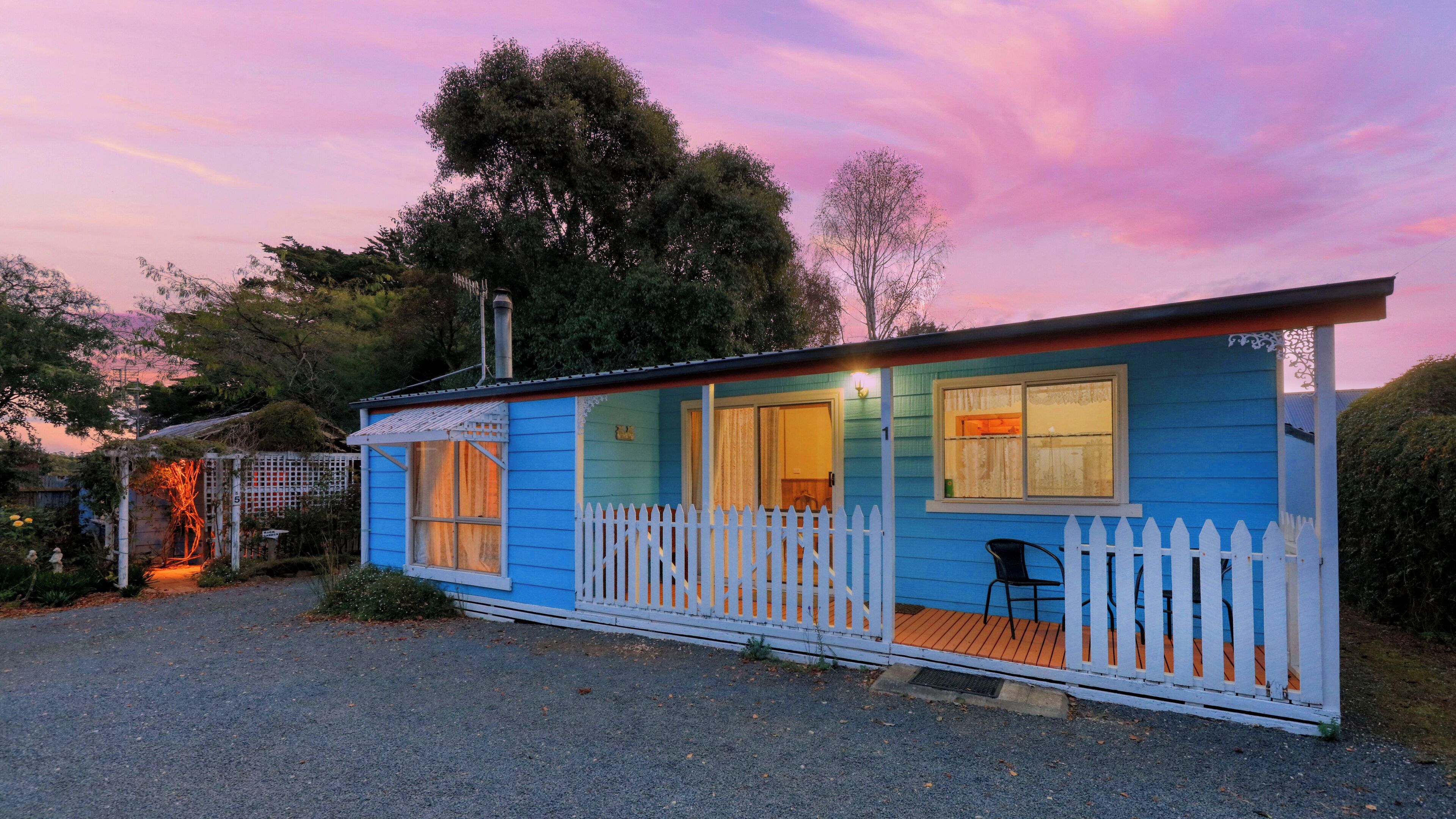 A small, single-story blue cabin with a white picket fence, a porch, and a chimney.