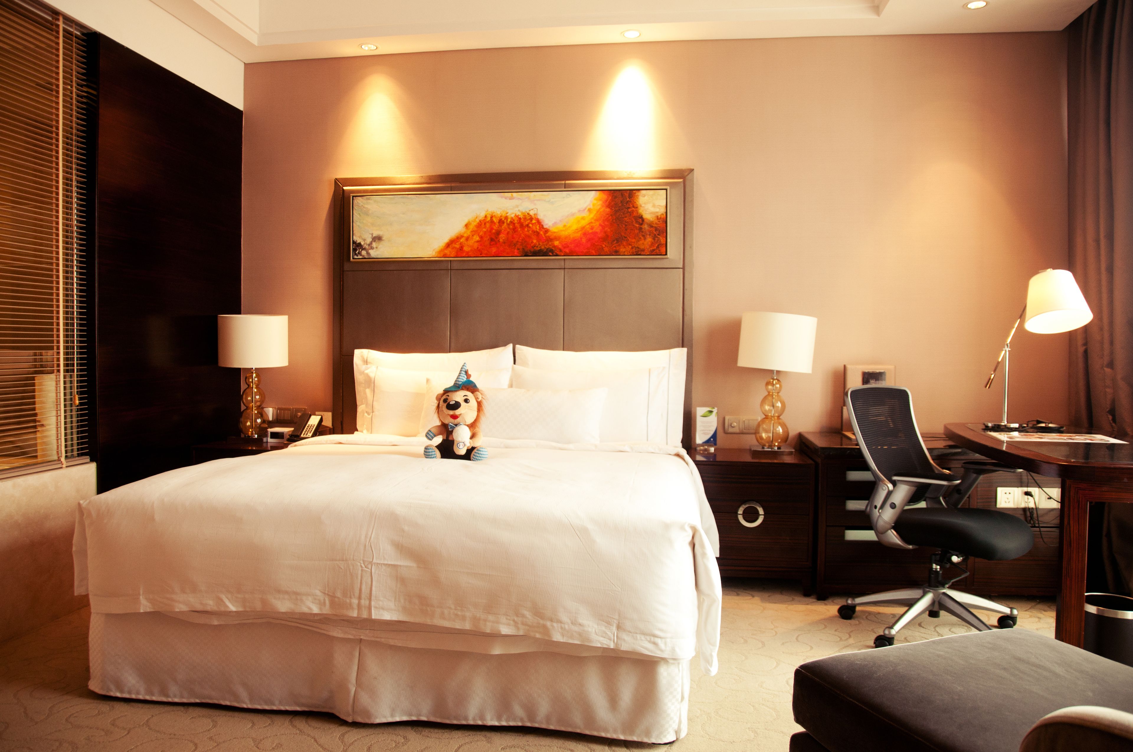 A hotel room with a large bed, two bedside tables with lamps, a desk with a chair, and a painting above the bed.