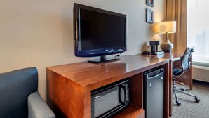 A hotel room with a flat-screen TV on a wooden console, a microwave, a coffee maker, and a desk with a chair.