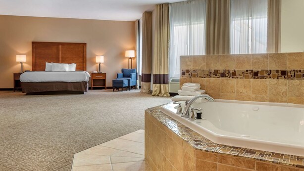 A hotel room with a large bed, a separate sitting area with a chair and table, a bathroom with a bathtub, and a window with curtains.