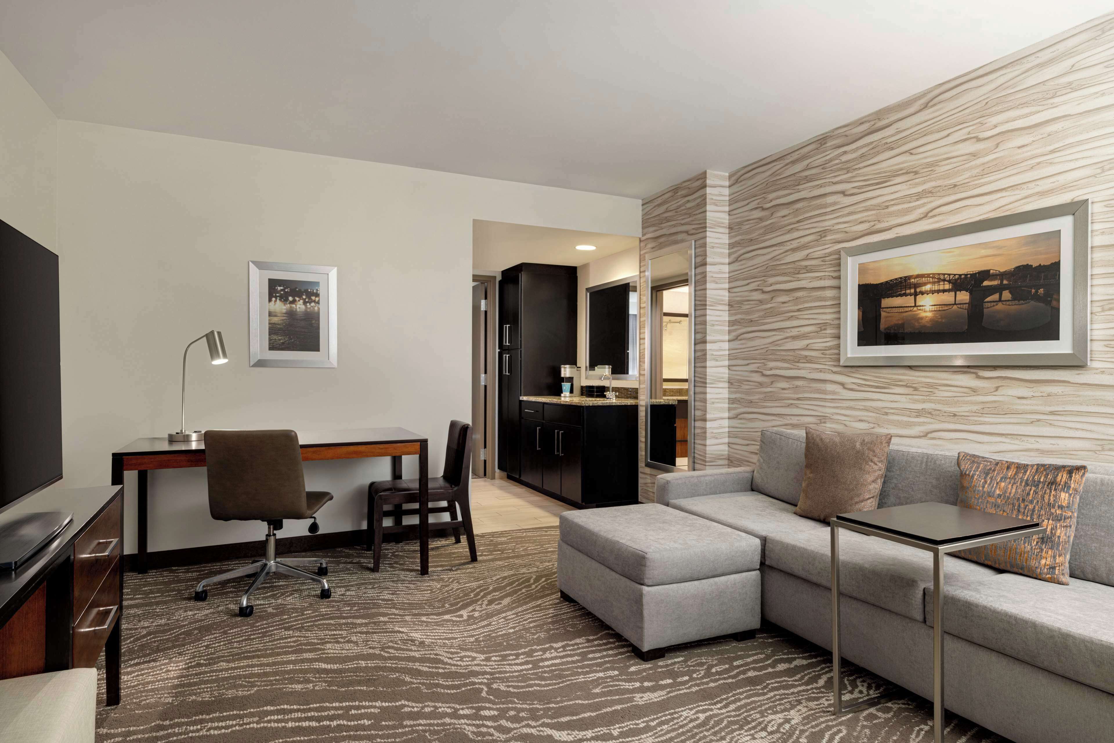 A modern hotel room with a grey sectional sofa, a small table, a desk with a chair, a flat-screen TV, and a framed picture on the wall.