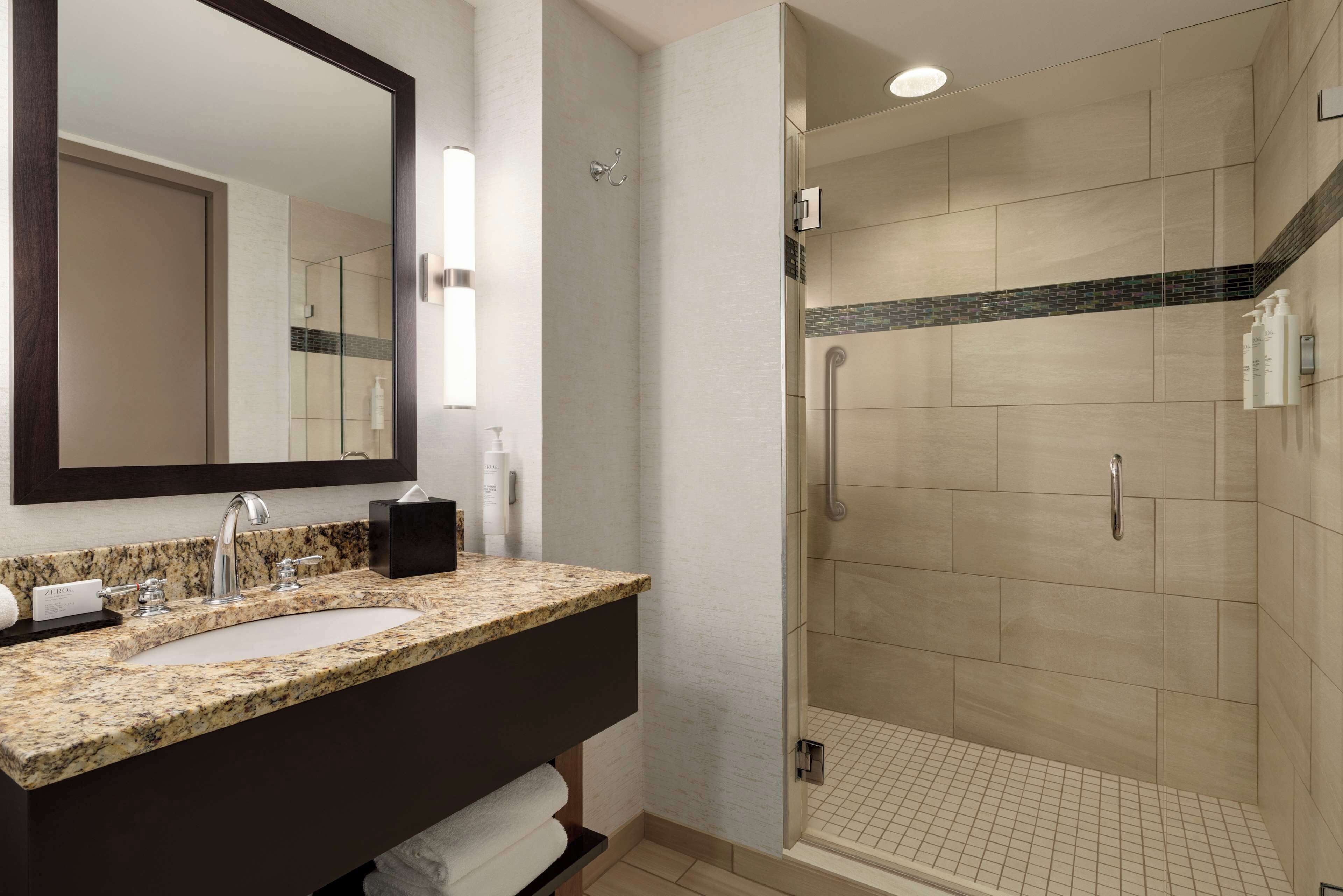 A bathroom with a granite countertop sink, a large mirror, and a walk-in shower with beige tiles.