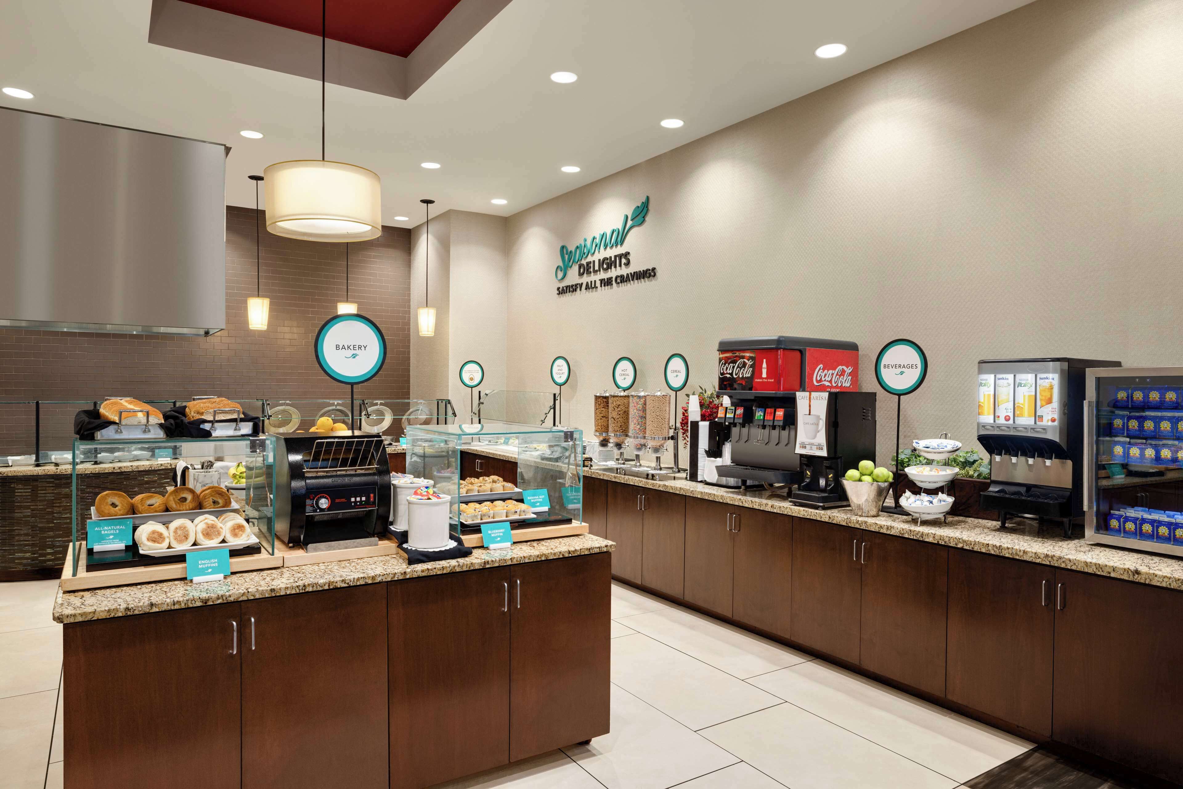 A hotel breakfast area with a bakery display, coffee machines, and beverage dispensers.