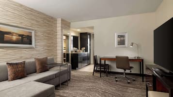 Une chambre d’hôtel moderne avec un canapé gris, une table à manger avec des chaises, un téléviseur à écran plat et un coin cuisine visible à travers une porte ouverte.