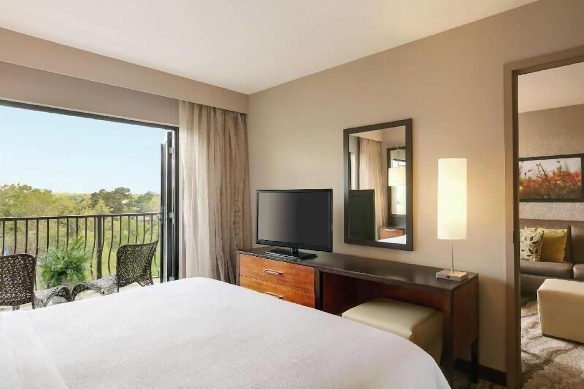 A hotel room with a bed, a television, a desk with a lamp, a mirror, and a balcony with chairs.