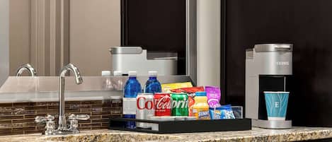 A hotel bathroom with a granite countertop, a sink with a modern faucet, a coffee maker, and a tray with various beverages and toiletries.