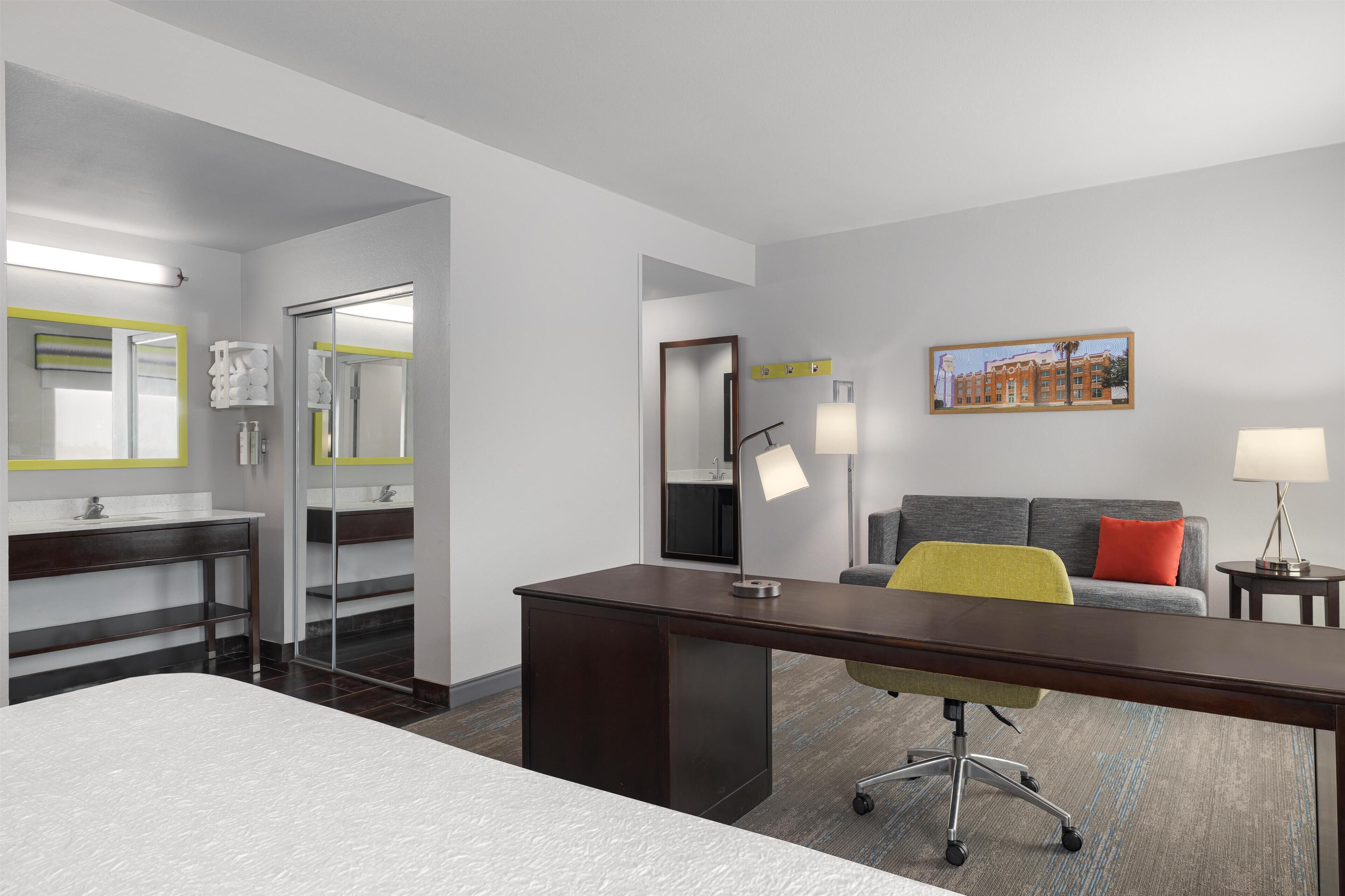 Habitación de hotel con cama, escritorio, sillón, sofá, baño con espejo y un cuadro en la pared.