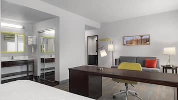 Habitación de hotel con cama, escritorio, sillón, sofá, baño con espejo y un cuadro en la pared.
