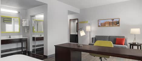 A hotel room with a bed, a desk, a chair, a sofa, a bathroom with a mirror, and a painting on the wall.