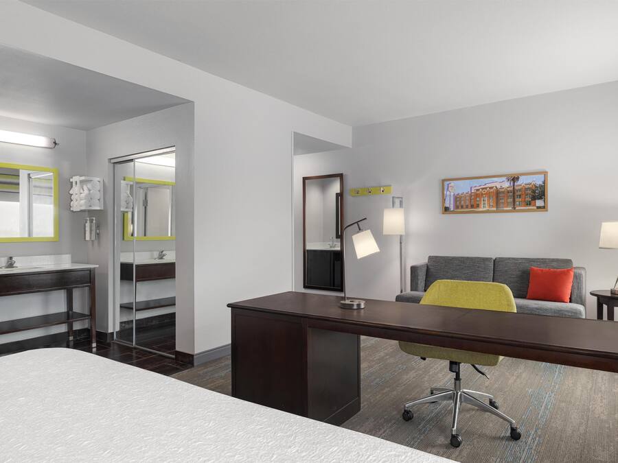 A hotel room with a bed, a desk, a chair, a sofa, a bathroom with a mirror, and a painting on the wall.