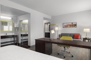 Habitación de hotel con cama, escritorio, sillón, sofá, baño con espejo y un cuadro en la pared.
