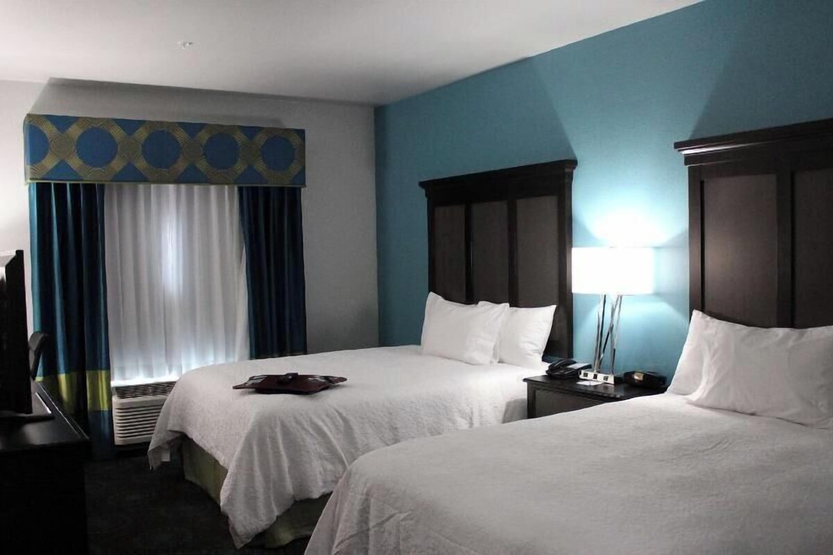 Habitación de hotel con dos camas, un escritorio, una silla, una lámpara y una ventana con cortinas.