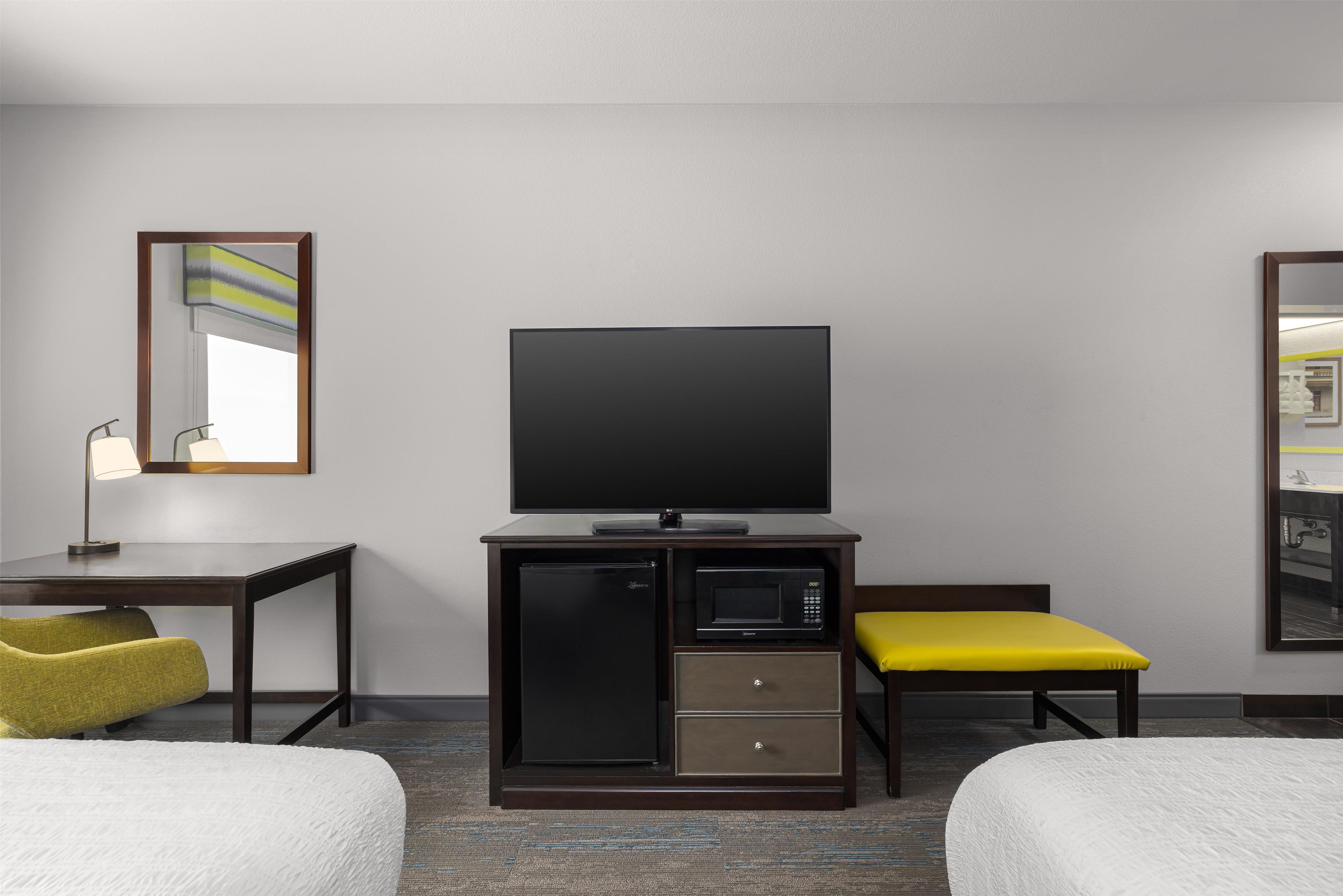 A hotel room with a flat-screen TV, a minibar, a desk with a lamp, two beds, and a mirror.