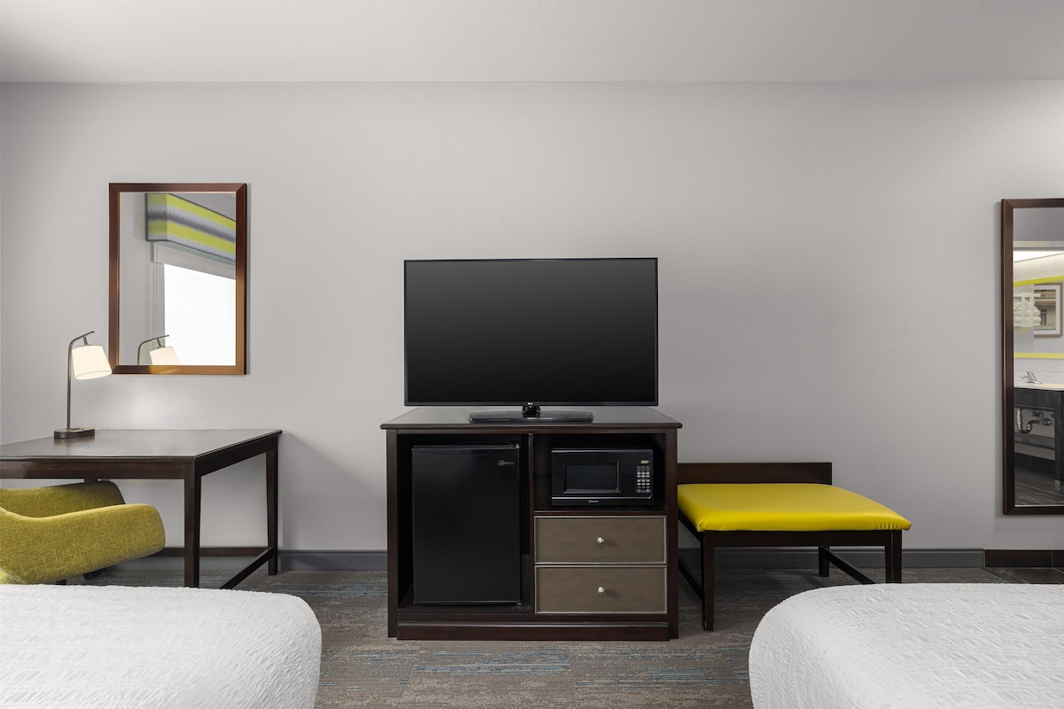 A hotel room with a flat-screen TV, a minibar, a desk with a lamp, two beds, and a mirror.