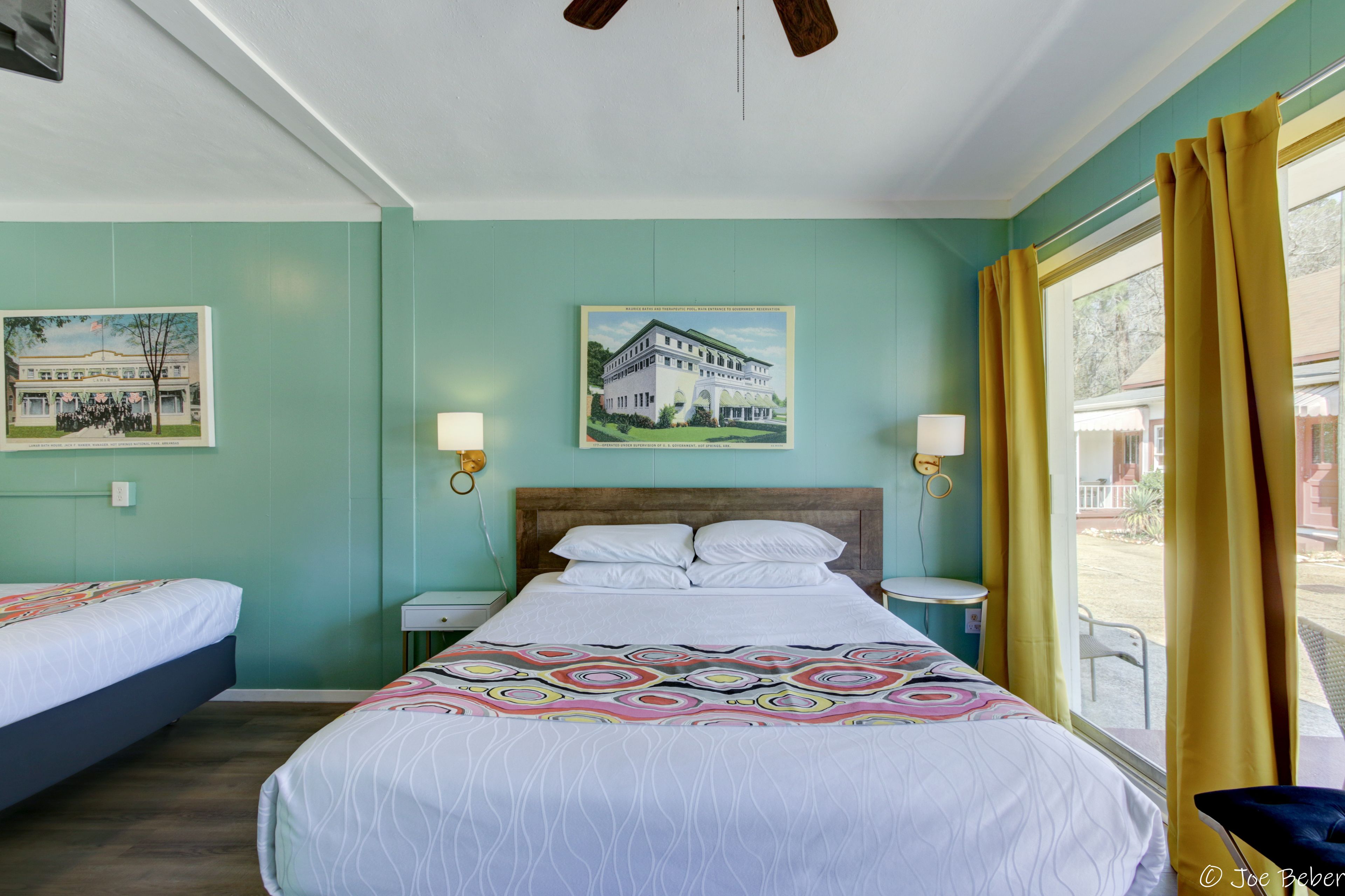 Una habitación de hotel con una cama, dos mesitas de noche, un ventilador de techo y una ventana con cortinas.