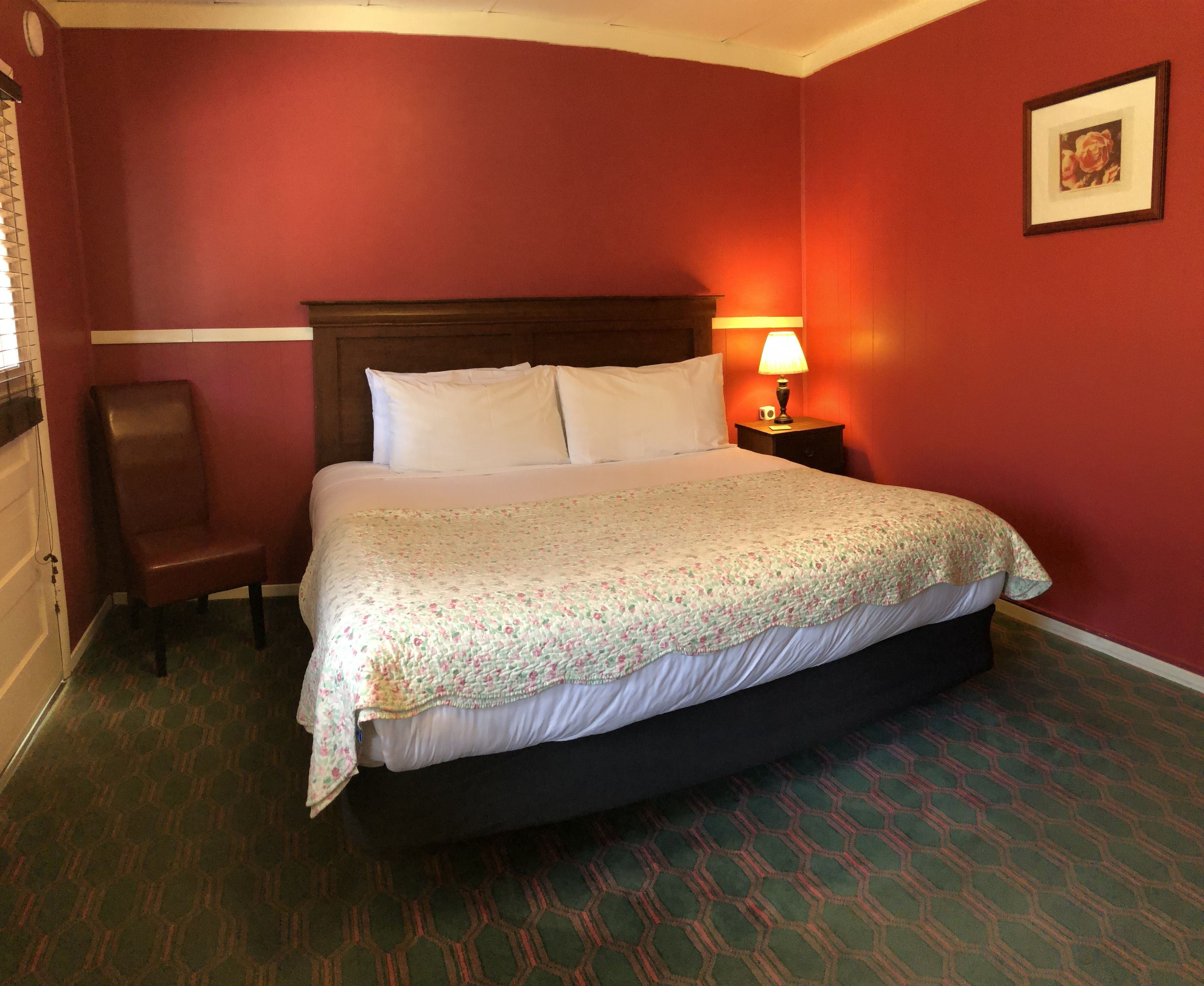 Una habitació d'hotel amb un llit gran, un panell de fusta al capçal, una tauleta de nit amb una làmpada, una cadira i un quadre encimbellat a la paret.