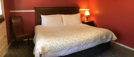 A hotel room with a large bed, a wooden headboard, a nightstand with a lamp, a chair, and a framed picture on the wall.