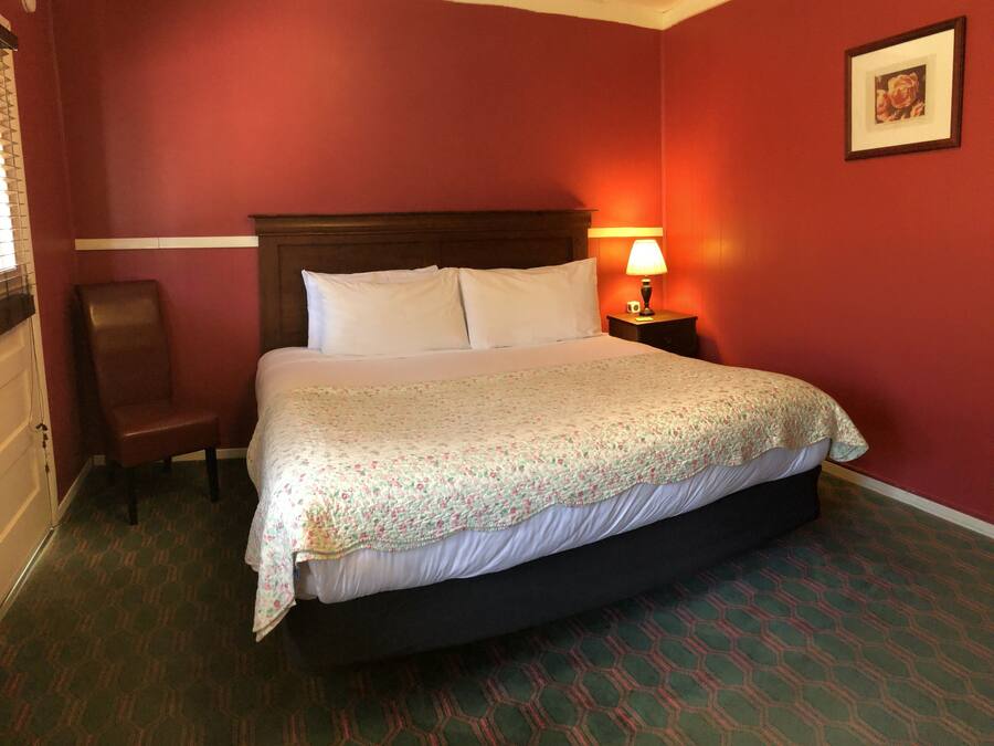Una habitació d'hotel amb un llit gran, un panell de fusta al capçal, una tauleta de nit amb una làmpada, una cadira i un quadre encimbellat a la paret.