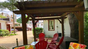 Een overdekt terras met rode stoelen en een tafel, een barbecue en een pergola met lichtslingers.