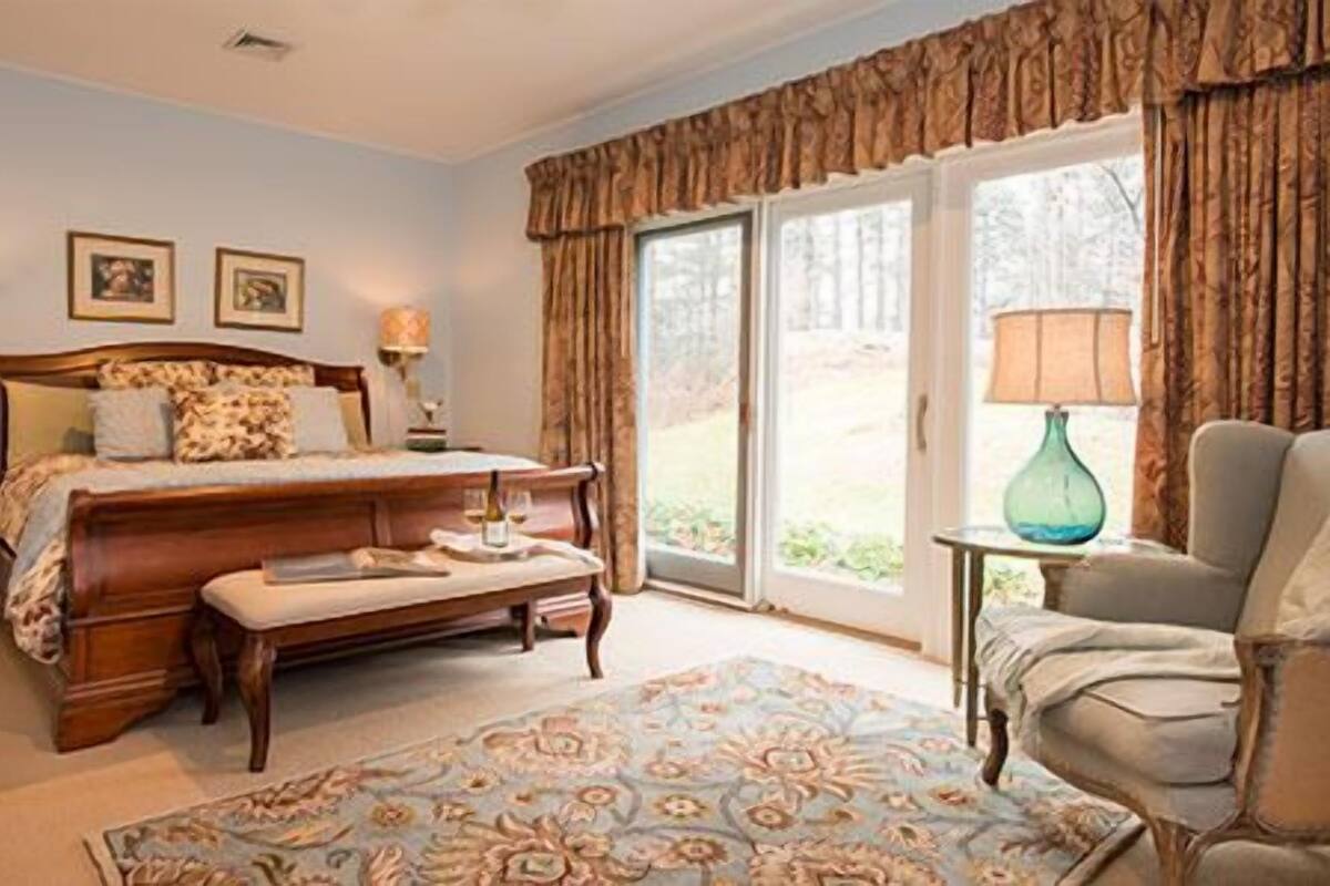 Un dormitorio con una cama grande, una mesita de noche, una silla, una ventana con cortinas y vistas al exterior.