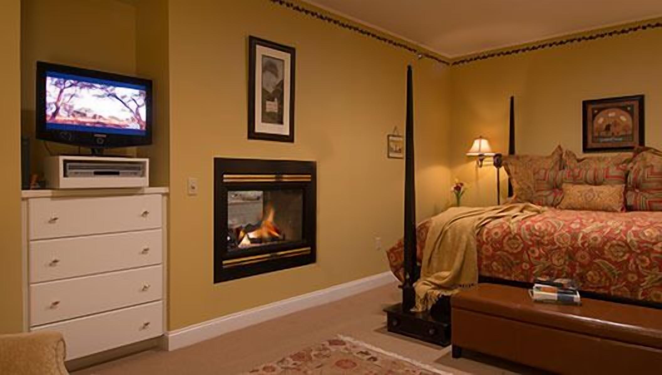 A bedroom with a fireplace, a television, a bed with patterned bedding, and a nightstand with a lamp.