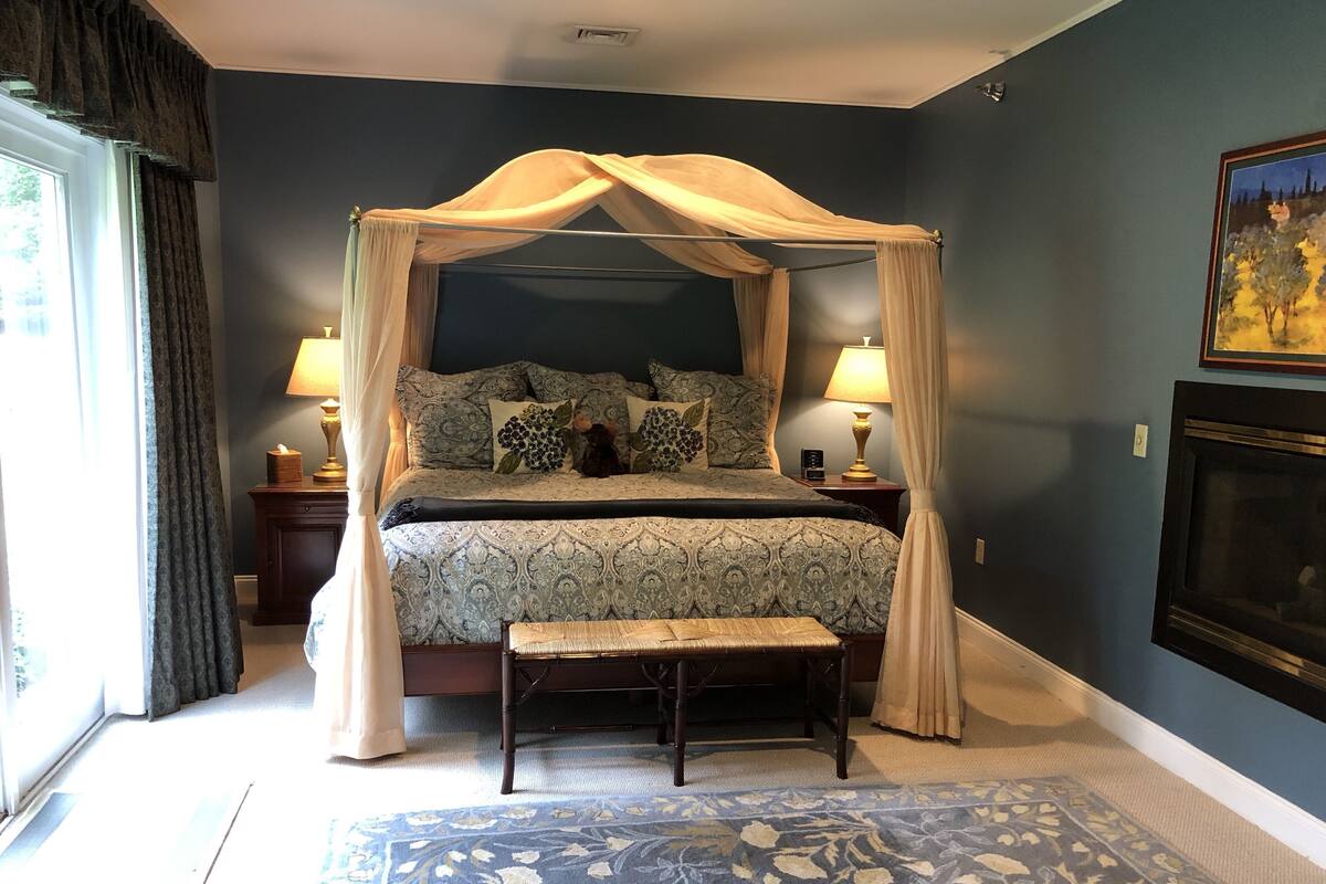 Un dormitorio con cama con dosel, mesita de noche con lámpara, y un cuadro grande en la pared.
