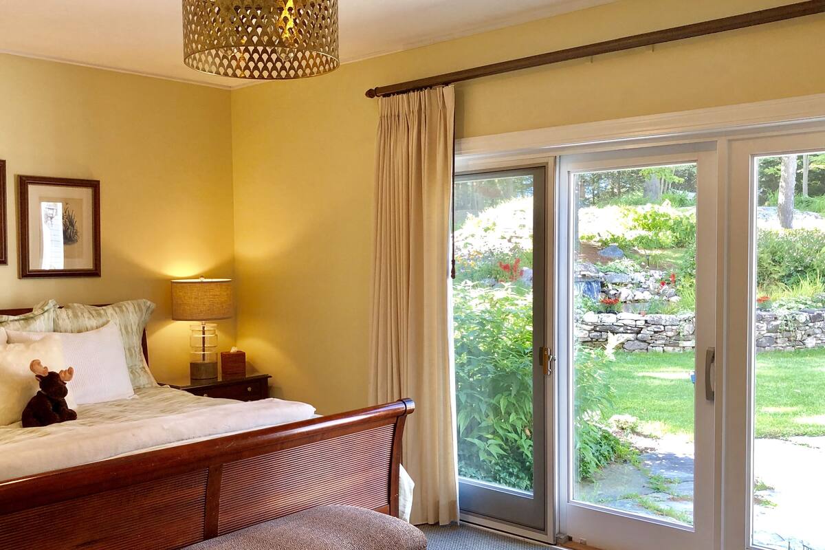 Un dormitorio con cama, mesita de noche con lámpara, ventana con cortinas y vista a un jardín.