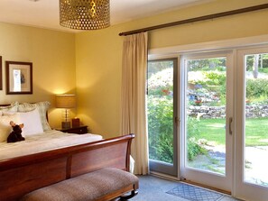 A bedroom with a bed, a bedside table with a lamp, a window with curtains, and a view of a garden.