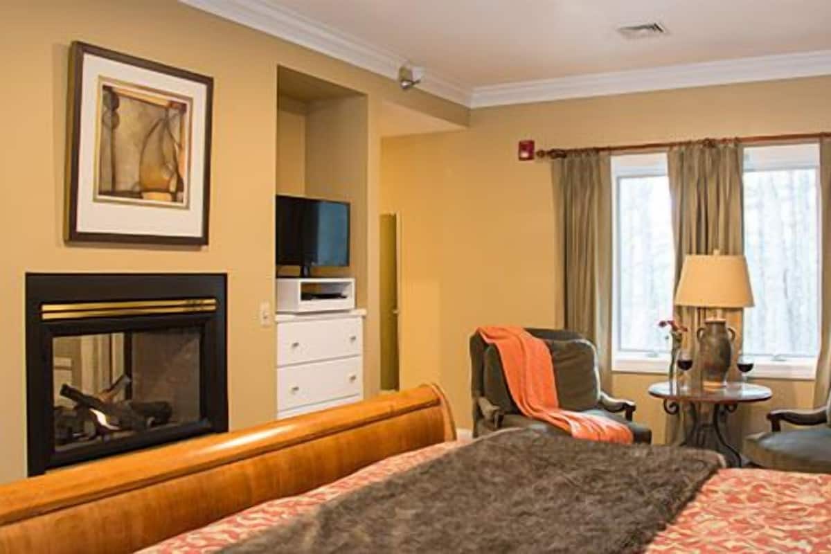 Habitación de hotel con cama, dos sillones, chimenea, televisor y ventana con cortinas.