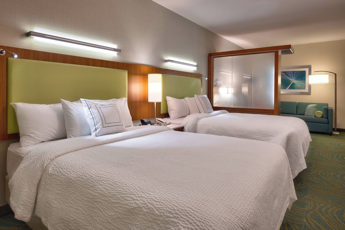 A hotel room with two beds, a green headboard, a bedside table with a lamp, and a blue sofa in the corner.