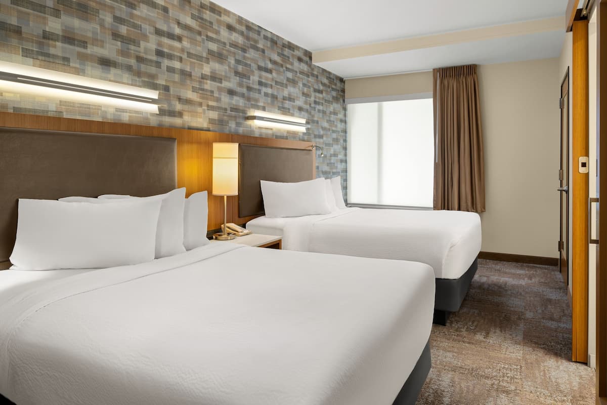 A hotel room with two beds, a headboard with integrated lighting, a bedside table with a lamp, and a large window with curtains.