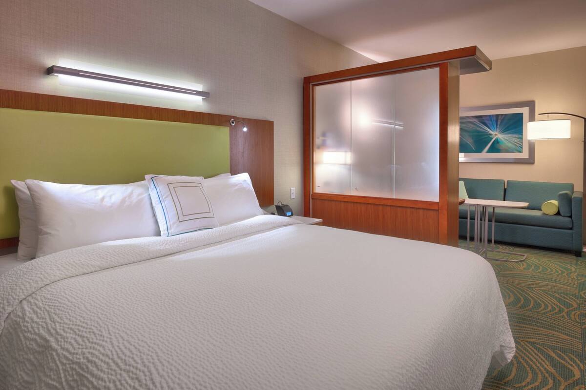 A hotel room with a large bed, a green headboard, a glass partition, a small table, a sofa, and a lamp.