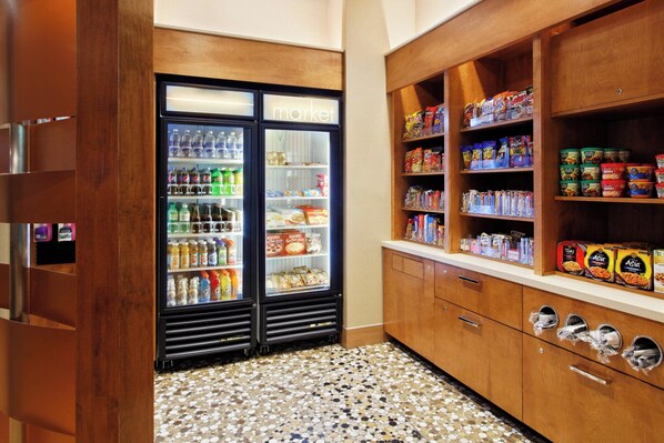 Uma cozinha compacta com geladeira repleta de bebidas e lanches, armários de madeira com alimentos e piso cerâmico.