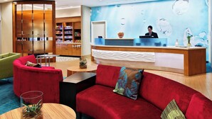 A hotel reception area with a modern design, including a reception desk, a red sofa, a green armchair, and a wooden bookshelf. - SpringHill Suites Pensacola (Pensacola)