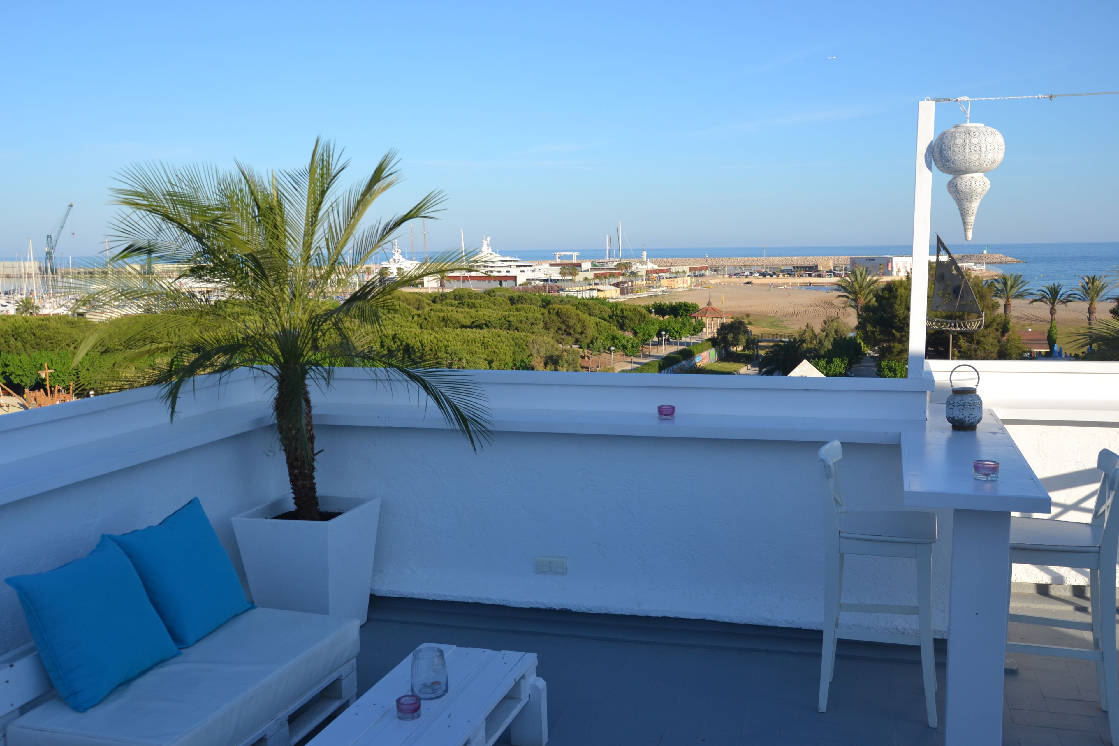 Terraza en la azotea con un sofá blanco, una mesa y sillas. Se ve la playa y barcos al fondo.