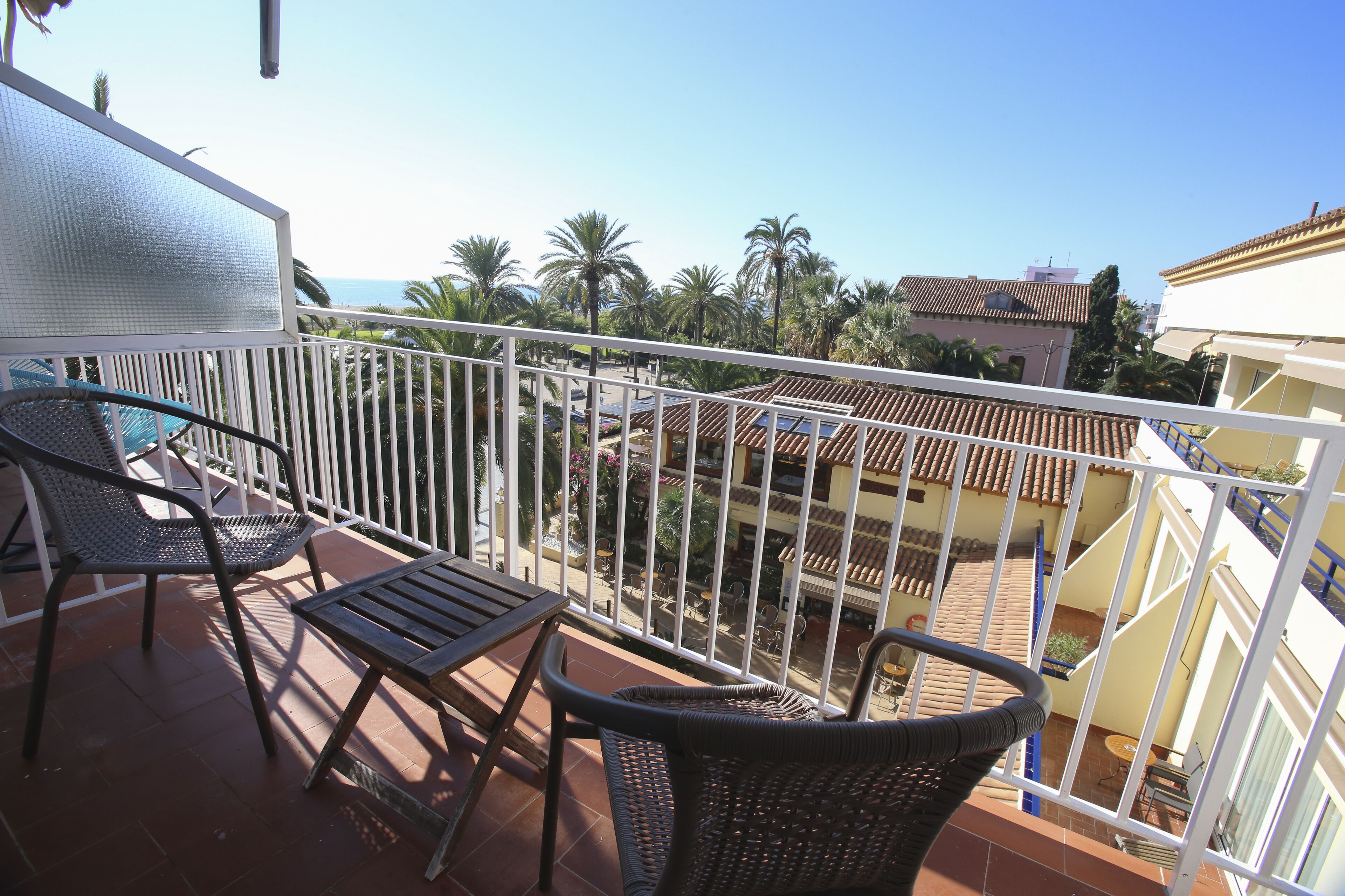 A balcony with wicker furniture, a view of palm trees, and a tiled roof.