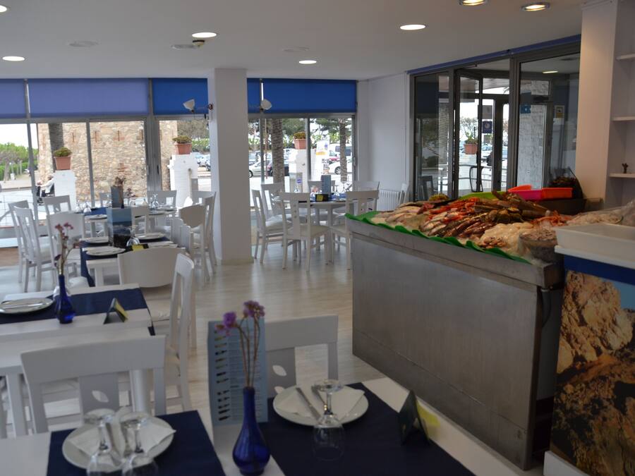 Un restaurante con mesas y sillas blancas, una barra con exhibición de mariscos y amplias ventanas con vista a la calle.