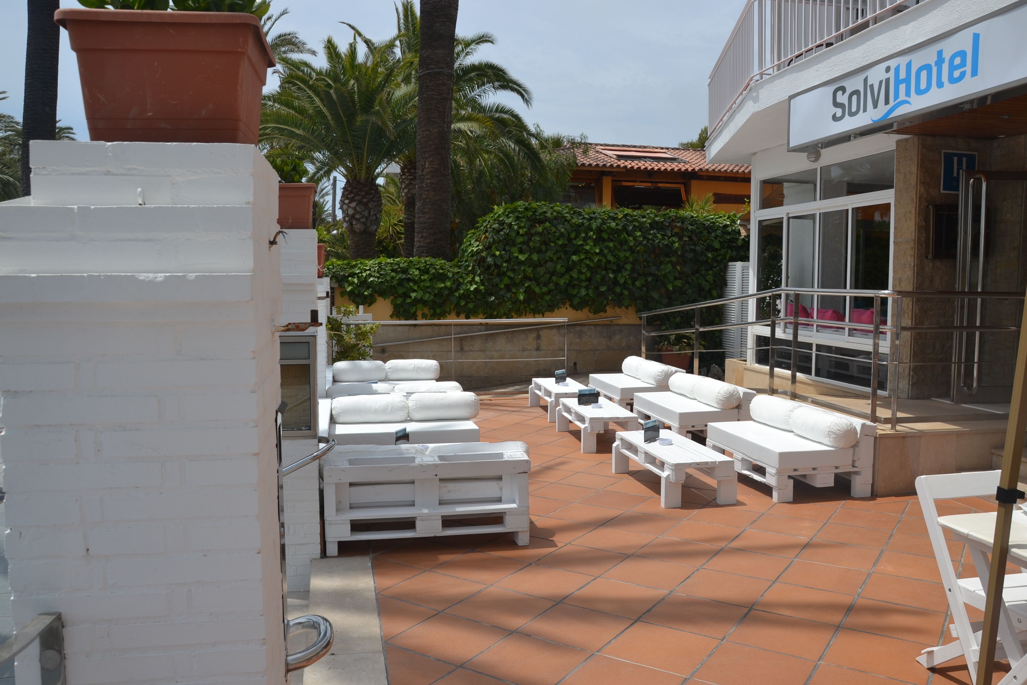 Un área de estar al aire libre con mobiliario blanco, piso de baldosas y un edificio con la leyenda "SolviHotel" al fondo.