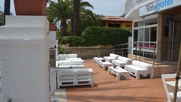 Un área de estar al aire libre con mobiliario blanco, piso de baldosas y un edificio con la leyenda "SolviHotel" al fondo.