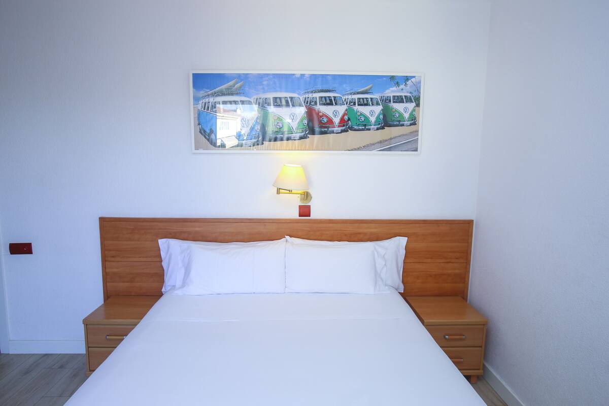 Habitación de hotel con cabecera de madera, cama con ropa blanca, mesita de noche con cajones, lámpara de pared y un cuadro con autobuses de colores en la pared.